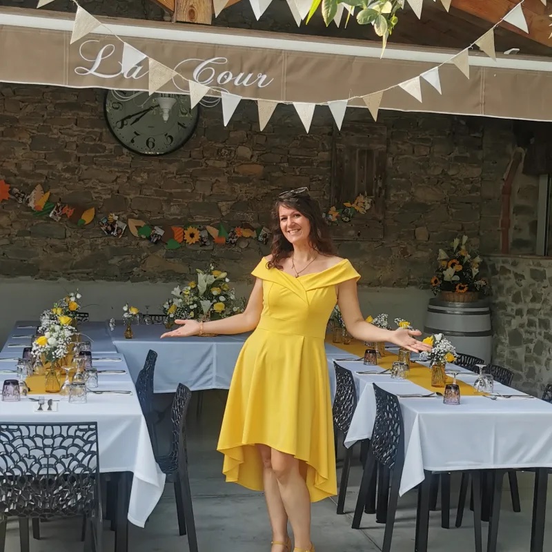 Claire Lys, en robe jaune et bras ouverts, présente en souriant une table d'événement décorée aux tournesols dans une cour en pierre