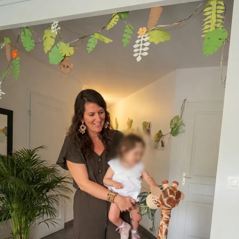 Maman tenant son bébé dans un décor de baptême sur le thème de la jungle : guirlande de feuillage et girafe en peluche