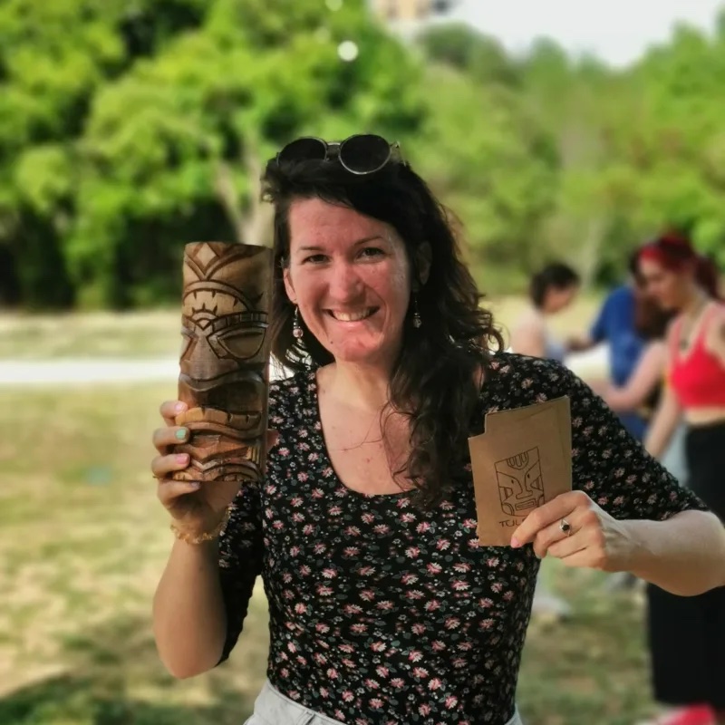 Claire Lys souriante lors d'une animation de jeu en plein air, un totem sculpté et une carte de jeu en main